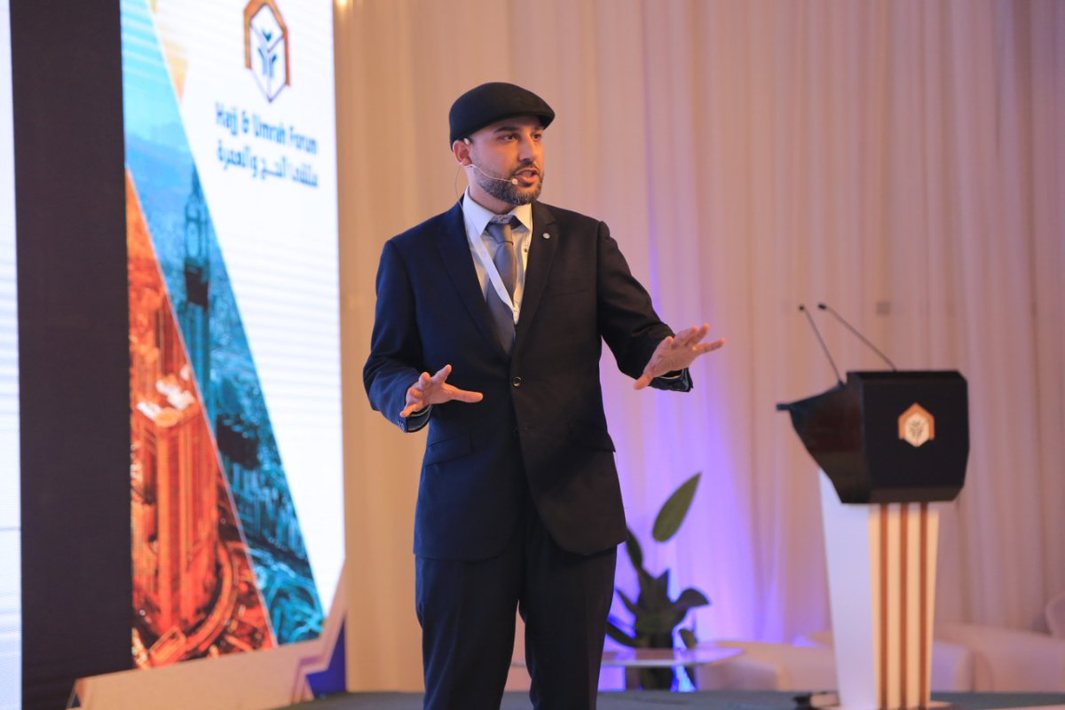 Mohsin Tutla presenting on stage at the Hajj and Umrah Forum, gesturing with both hands