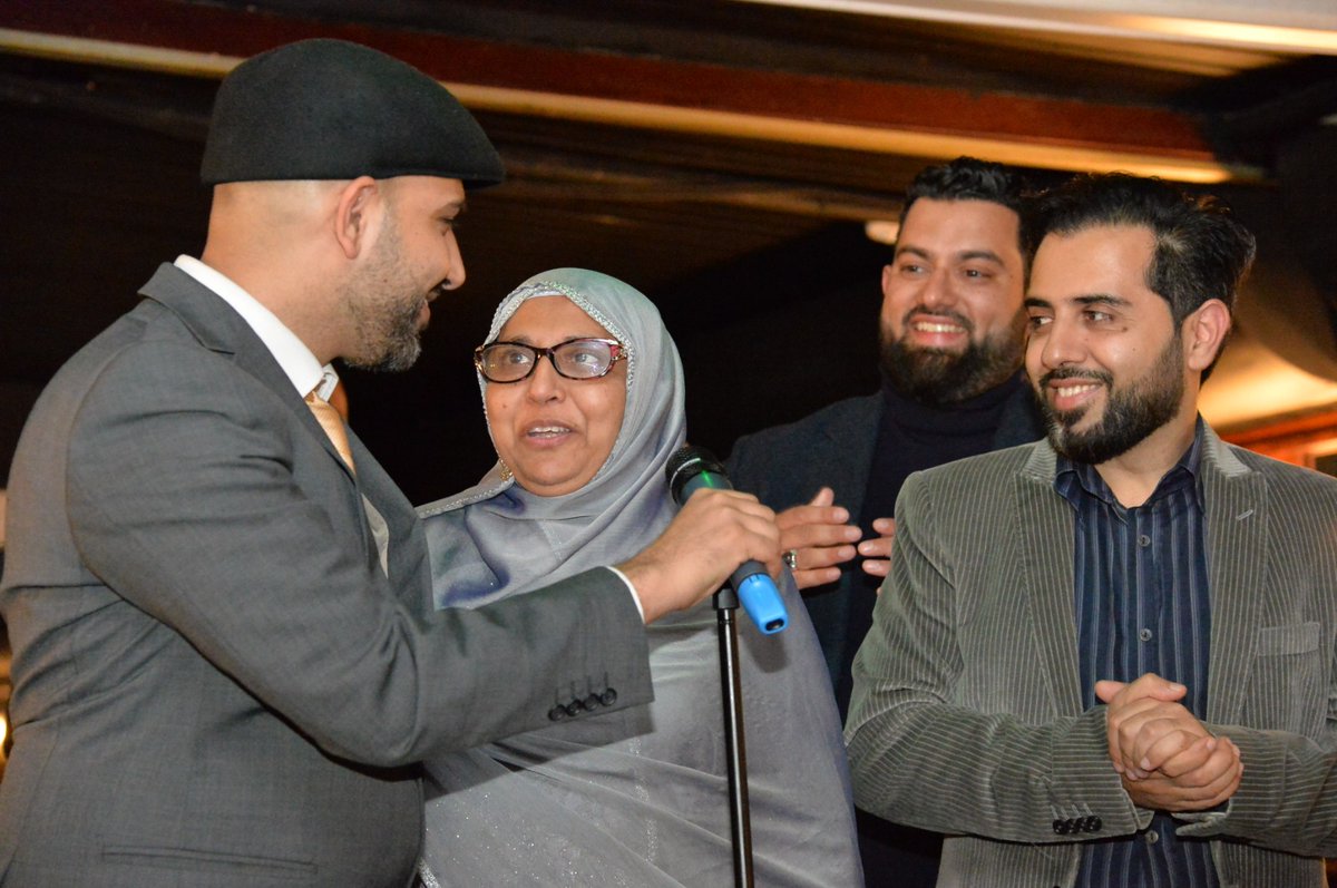 Mohsin Tutla holding the microphone for his mother on stage at an evening event, with guests smiling beside them